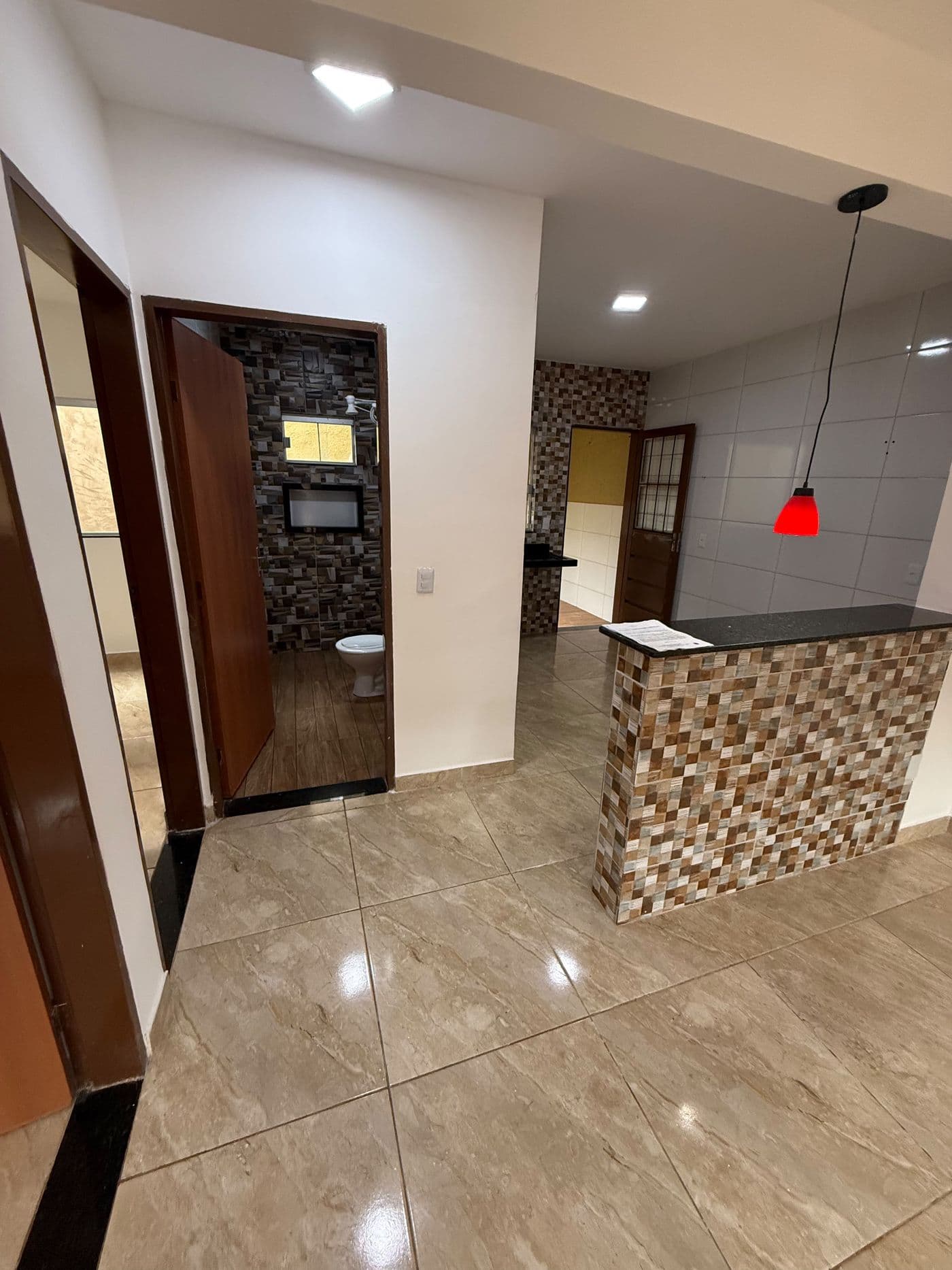 Sala ampla com piso porcelanato, bancada de granito revestida em pastilha separando a cozinha e acesso ao banheiro.
