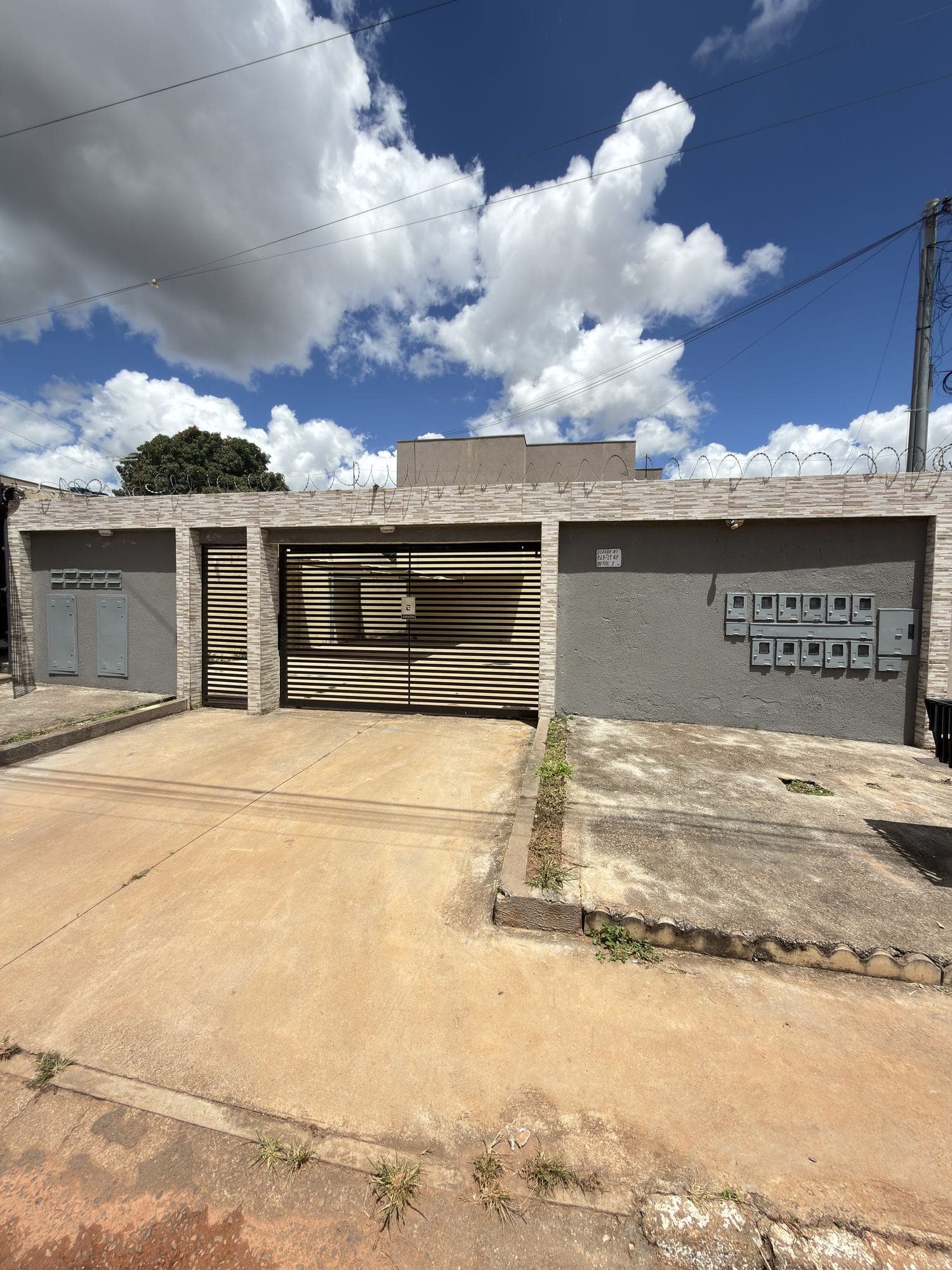 Fachada do Residencial Lopes V: muro em grafiato cinza com faixa de pedra clara, portão de garagem em lâminas cor de areia e medidores à direita.